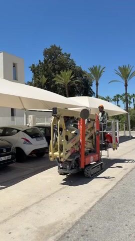 A person wearing a hard hat and safety vest operates a compact tracked aerial lift. The lift is orange and yellow, with a basket at the end of its articulated arm. The person is sitting in the basket, holding onto the controls. The lift is positioned beneath a large white awning, which is supported by poles. In the background, there are several cars parked, a modern white building, and palm trees. The sky is clear and blue. The overall scene suggests outdoor maintenance or construction work being performed on a sunny day.