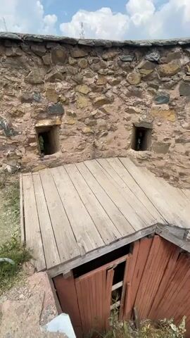 This media file captures a view from within an ancient stone structure, likely a fort or castle, overlooking a valley and a town. The foreground is dominated by a rustic wooden walkway and stairs, some of which appear worn and in need of repair. The stone wall of the structure features several small, square openings, characteristic of embrasures or loopholes, suggesting a defensive purpose. Beyond the wall, a panoramic view of a lush green valley with scattered buildings and a distant town is visible under a bright, cloudy sky. The overall impression is one of historical significance and a serene, albeit slightly neglected, natural setting. The time of day appears to be daytime, with good natural light, and the weather is fair with scattered clouds. The location cues, such as the architecture and the landscape, strongly suggest Srinagar, India. No individuals are visible in the frame.