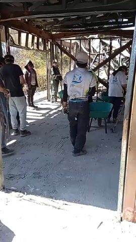 Certainly! Here's a detailed description of the video:

The video shows a group of people inside a partially destroyed building. The structure appears to be made of metal framing, with significant damage to the roof and walls, possibly from a fire or other disaster.

Several individuals are wearing white T-shirts with a logo, while others are in casual clothing. Some people are using shovels to clear debris, and wheelbarrows are present to transport materials. The floor is covered with rubble and ash. The video seems to be documenting a cleanup effort, possibly involving volunteers.