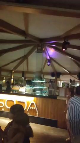 The video shows a lively indoor venue, possibly a bar or restaurant, with a wooden ceiling and exposed beams. People are seen mingling and dancing. In the foreground, a wooden counter with a glass display case is visible, suggesting a bar area. There are spotlights illuminating the space, and some areas are bathed in purple light. In the background, a stage or dance floor can be seen with more people gathered. The overall atmosphere appears festive and social.