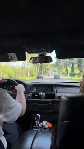 The video shows the interior of a car in motion, filmed from the back seat. A person with fair skin and short brown hair is driving. The person is wearing a white shirt. The car appears to be a BMW, judging by the interior. The car is traveling on a road lined with trees on both sides. There are several other cars visible ahead on the road. The sun is shining. The radio is playing pop music.