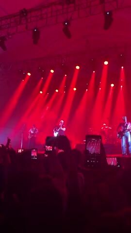 The video shows a band performing on a stage bathed in red light. The stage is set up with several spotlights creating beams that shine down on the band members. From left to right, there is a guitarist, the lead singer, a drummer in the background, and another guitarist on the right side of the stage. The lead singer is in the center, holding a microphone and singing. Below the stage is a crowd of people, many of whom are holding up their phones to record the performance. The structural rigging of the stage is visible above, adding to the concert atmosphere.