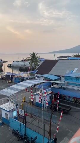 The video shows a ferry terminal at sunset. The sky is a soft orange and pink, with some clouds. In the distance, there are several ferries docked and at sea. In the foreground, there is a parking lot with cars and motorcycles. People can be seen walking around and working. The terminal buildings are blue and white, with striped awnings. There are also some red and white striped barriers. The overall scene is calm and peaceful.