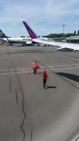 Here's a plain text description of the video:

The video shows an airport tarmac. Two workers wearing orange vests walk across the tarmac. One worker is carrying a piece of equipment and the other worker has red flags in his hand. In the background, there are two planes, one from Ryanair and one from Wizzair. The sky is blue with some clouds. A portion of an airplane wing is visible in the foreground. The video appears to be taken from inside an aircraft, looking out the window.