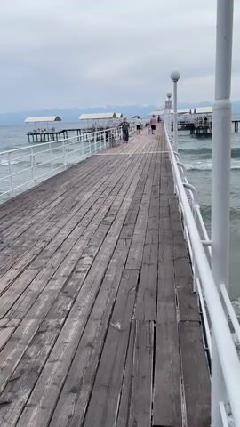 A wooden pier extends into a choppy, gray body of water. The pier has white railings and lamp posts. Several people walk on the pier. In the distance, there are structures on stilts over the water and mountains under a cloudy sky. The camera pans to the right, showing the water and a metal fence partially submerged in the waves near the shore. The waves are creating foam and are breaking against the shore.