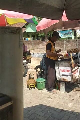 The video shows a street scene with several people and food stalls. In the foreground, a cylindrical object, possibly a pipe, is visible. To the left, colorful umbrellas provide shade over a food stall. Further back, a man wearing a yellow shirt and a cap is standing near a food cart labeled "TAKOKAYAK". Another man in a blue shirt is seen walking near a similar food cart. The background reveals buildings and trees, suggesting an outdoor urban or suburban environment. The ground is paved with bricks. The video appears to be filmed from a handheld device, with a slightly tilted perspective.