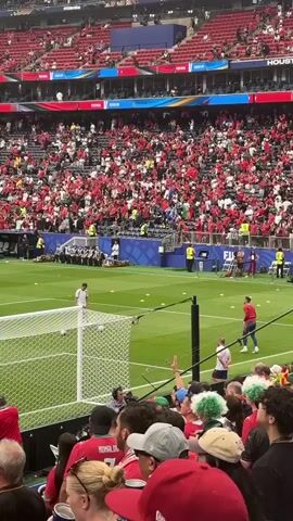 The video shows a crowd at a soccer game. The spectators are wearing red and white clothing. The field is green, and there is a soccer goal in the foreground. The crowd is cheering and waving their arms. The stands are filled with people. Some people are wearing hats and scarves. The sky is not visible. The video is shot from a high angle, looking down at the field and the crowd.
