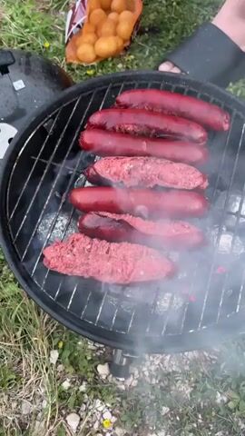A close-up aerial view shows a small charcoal grill with several sausages and hamburger patties cooking on the grate. Some of the patties are split open, revealing raw meat. A pair of tongs is used to flip one of the patties. Steam rises from the food and the grill. In the background, out of focus, is a bag of small potatoes and a person's foot wearing a black sandal. The grill is set on a grassy area with some small yellow flowers visible. The video is shot from a high angle, looking down at the grill.