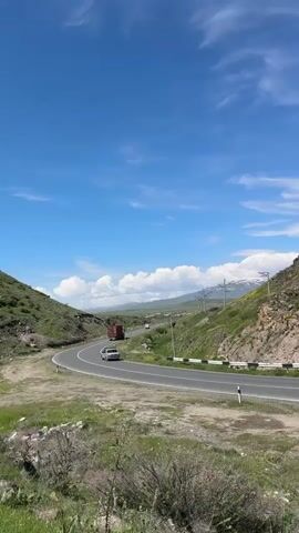 The video shows a scenic view of a winding road through green hills under a bright blue sky with scattered clouds. A few cars and a red truck are visible on the road. Power lines run along the side of the road. The landscape is grassy with some rocky areas. The shot appears to be taken from a slightly elevated position.