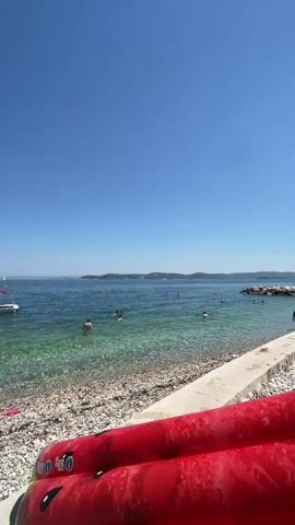 The video displays a sun-drenched beach scene on a clear, sunny day. The sky is a vibrant, cloudless blue, meeting the calm, turquoise water. The foreground is dominated by a large, inflatable object designed to resemble a sliced watermelon, its bright red and black seeds clearly visible. The beach itself is composed of small, light-colored pebbles. In the shallow, clear water, several people are swimming and enjoying themselves. Some are wading, while others are further out, swimming. A white boat with a red trim is visible in the middle distance, and a few other people are scattered on the beach and in the water further along the coastline. The water clarity allows for a view of the pebbly seabed in the shallower areas. The overall impression is one of a leisurely day at the beach.