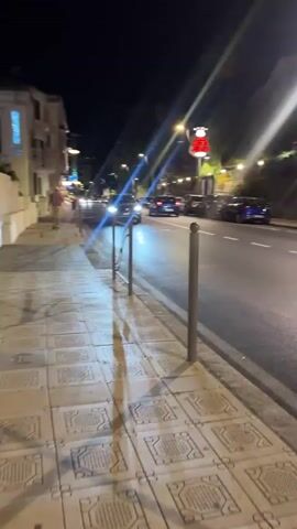 The video pans rightward along a sidewalk at night. The sidewalk is paved with square tiles featuring a decorative pattern. A metal railing runs along the edge of the sidewalk, separating it from the street. On the left side of the street, there are buildings. A few people can be seen walking on the sidewalk. The street itself is dark but illuminated by streetlights and the headlights of passing vehicles. Several cars are parked along the right side of the street. A white bus is traveling down the street, and its taillights are visible. In the background, there are trees and a hillside. A red sign with white lettering is visible hanging above the street. The overall atmosphere is that of a quiet urban street at night.