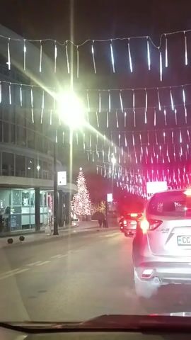 The video shows a nighttime street scene with Christmas decorations. The camera is likely mounted inside a car, looking through the windshield. The street is lined with buildings on the left and trees on the right. Above the street, strings of white icicle-shaped lights are hung. A large, brightly lit Christmas tree is visible in the distance, with a star on top. Several cars are on the street, including a silver Mazda CX-5 in front of the camera car, with its brake lights illuminated. There are a couple of people walking near the Christmas tree. A street lamp is on the left, shining brightly.