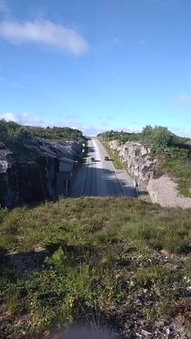 The video shows a view from a grassy hill looking down onto a paved road that goes uphill and curves to the right. The road is flanked on both sides by steep, rocky cliffs. There are a few cars on the road, and the sky is blue with some white clouds. There appears to be a small building or structure on the left side of the road, built into the cliff face. The area is sparsely vegetated with grass and some bushes. The lighting suggests it is daytime, likely with sunny weather.