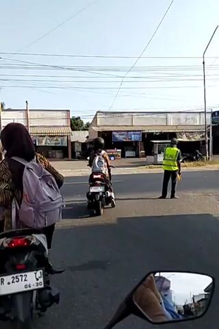 The video shows a street scene with several people on motorbikes. In the foreground, a motorbike with a license plate starting with "R 2572" is visible. A person wearing a headscarf and a backpack is riding it. In the middle ground, another motorbike with a person wearing a helmet and a backpack is seen. Further in the background, there are several shops and buildings lining the street. The sky is clear and the day appears to be sunny. The overall scene depicts a casual street setting with pedestrian and vehicle traffic.