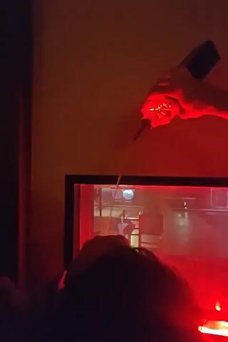 A first-person perspective video shows a person holding a clear plastic bottle with a metal spout over a large flat-screen television. Red light emanates from the television and the surrounding environment. The person's hand is visible as they extend the bottle towards the screen. The video then shifts to show the back of a person's head and shoulders, with red light reflecting on their hair. Further into the video, another person's head and shoulders are visible in the foreground, and a person with dark hair wearing glasses is partially visible in the middle ground, looking down and covering their mouth with their hands. People are visible on the television screen, which displays a scene that appears to be a crowded event with red lighting.