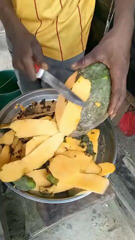 A man in a yellow and red striped shirt is peeling a green papaya with a knife over a large metal bowl. The bowl is filled with papaya peels and seeds. The man's hands are dark and he is holding the papaya firmly. The scene appears to be outdoors, with glimpses of green foliage and other objects in the background.