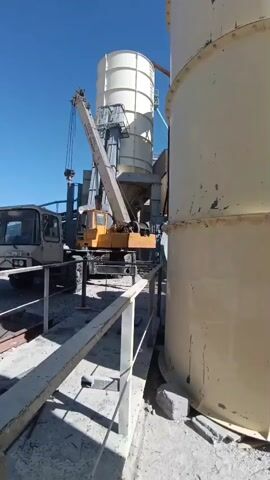 This media file captures an outdoor scene at what appears to be an industrial site, possibly a construction or processing facility, in Aragatsavan, Armenia. The time of day is daytime, indicated by the bright sunlight and clear blue sky, suggesting fair weather.

Dominating the frame are two large, cylindrical structures, likely silos or storage tanks, one predominantly light yellow and another lighter in color, situated behind. A mobile crane, painted orange and yellow, is positioned between the silos, its boom extended and a load suspended from its hook. The crane appears to be actively engaged in some form of lifting or placement operation, suggesting ongoing construction or maintenance.

To the left of the crane is the cabin of a truck, also light-colored, with visible license plates. The surrounding ground is rough and appears to be a mix of gravel and concrete. A metal railing runs in the foreground, indicating a walkway or platform.

While no people are clearly visible in the frame, their presence is implied by the operation of the crane and truck. The overall atmosphere is one of industrial activity and work. The image does not convey specific emotions, but rather a sense of purposeful action within an industrial setting. There is text visible on the side of the crane, but it is too small and blurry to be legible. The license plate on the truck also has text, but it is likewise unreadable. The presence of industrial silos and a crane points towards an environment involved in the storage or processing of materials, possibly related to agriculture, construction, or manufacturing.