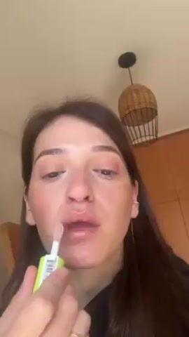 A woman is applying lip gloss in a close-up video. She has brown hair, fair skin, and is wearing minimal makeup. She is holding a lip gloss tube with a green and white label and a clear applicator. The room is lit by natural light, and there is a wooden cabinet, a woven pendant light, and part of a wooden chair visible in the background.