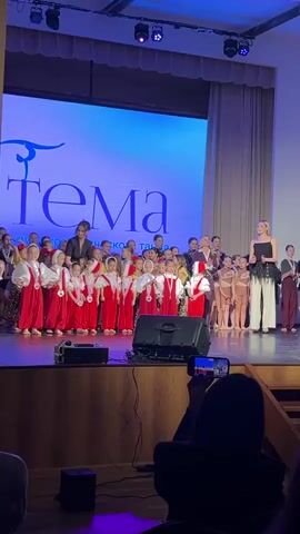 Here is a description of the video:

A group of young children, mostly girls, are standing on a stage in a performance setting. They are lined up and appear to be taking a bow or receiving recognition after a performance. The children are wearing different costumes; a large group of them is dressed in red and white traditional outfits.  

Several adult women stand among them, likely teachers or organizers. The women are wearing more formal attire. One woman is speaking, possibly giving a speech or announcing something.

The stage has a wooden surface. A large screen in the background displays text in a language that appears to be Russian. Stage lighting is visible above.

In the foreground, a person is recording the scene with a smartphone. The focus is on the stage and the performers. The atmosphere is celebratory.
