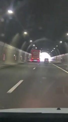 The video shows a view from inside a vehicle driving through a highway tunnel. The tunnel is illuminated by overhead lights. In front of the vehicle, a large orange truck with a white logo on its side and a white car are driving in the same direction. The tunnel appears to be heading towards an opening where daylight is visible, with trees and greenery visible outside. The camera is mounted low, suggesting it's capturing the view from the dashboard or front windshield.
