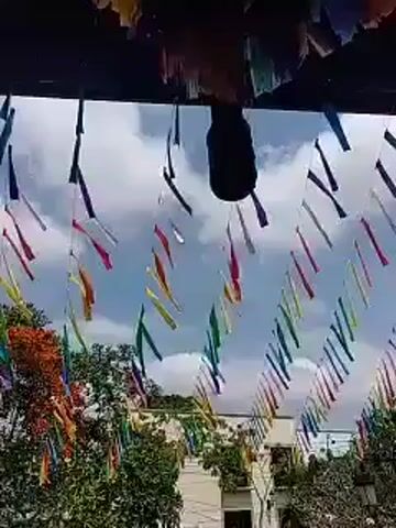 The video captures an outdoor scene in Xochitepec, Mexico, adorned with colorful decorations. Numerous brightly colored streamers or banners, appearing in a rainbow of hues, are suspended overhead, forming a festive canopy. They sway gently in the breeze against a bright, partially cloudy sky. A dark, cylindrical object, possibly a speaker or light fixture, hangs centrally among the decorations. Below the overhead canopy, the tops of green trees are visible, suggesting a lively street or public square. The scene is brightly lit by natural daylight, indicating a daytime setting. The vibrant decorations evoke a festive and celebratory atmosphere, likely for a local event or festival. No people or text are visible.