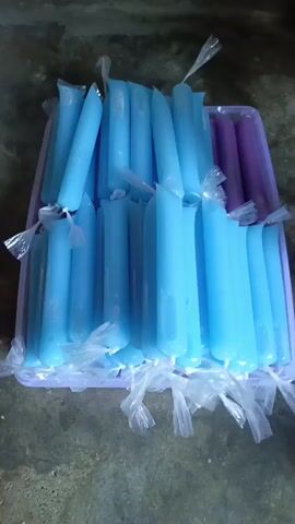 The video shows a close-up, slightly tilted shot of a purple plastic tray filled with many small, plastic-wrapped popsicles. The majority of the popsicles are a light blue color, and there are a few purple ones visible on the right side of the tray. The popsicles are arranged in several rows, standing upright. The lighting appears to be natural, coming from the top of the frame. The surface beneath the tray is rough and appears to be concrete or stone. The popsicles are sealed with clear plastic ties. There are no people or actions depicted in the video.