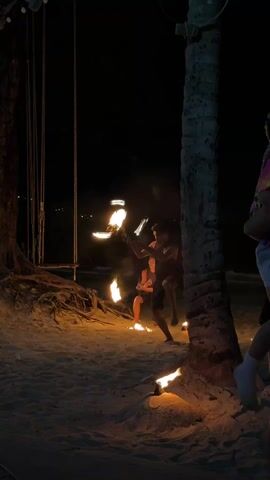 Two young men are performing a fire show on a sandy beach at night.  They are barefoot and wearing shorts and tank tops.  Each holds two flaming torches, and they are spinning and juggling them.  Fires are burning on the ground around them.  A rope swing is to the left, and a palm tree is to the right.  A person, likely a child, is partially visible in the foreground near the palm tree. Music plays in the background.