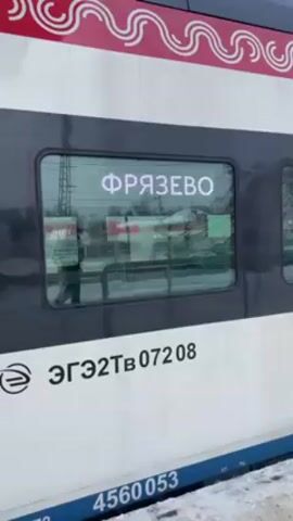 The video shows a train car parked at a station in what appears to be a snowy, cold environment. The train car is white with a gray stripe around the windows and a red decorative design at the top. The Cyrillic word "ФРЯЗЕВО" which transliterates to "FRYAZZEVO" is displayed in the window, indicating the train's destination or current location. Below the window, text is written in what appears to be Cyrillic, likely the train's identification number ЭГЭ2ТВ 07208. Below that, "4560053" is written in a blue strip. A person can be seen walking outside the train, reflected in the window. The ground is covered in snow.