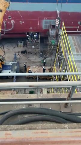The video shows a construction or repair site at a shipyard. Several people in white hard hats and work clothes are positioned around a metal platform on wheels, which is being lifted by a crane. Some are standing on scaffolding erected alongside the hull of a large ship, which is painted red on the bottom and white on the top. The crane is yellow and black and is labeled "MEGO-ACO". The camera is positioned above the scene, offering an overhead view. There are also various pieces of equipment and tools scattered around the site. The overall atmosphere suggests a busy and active industrial environment.
