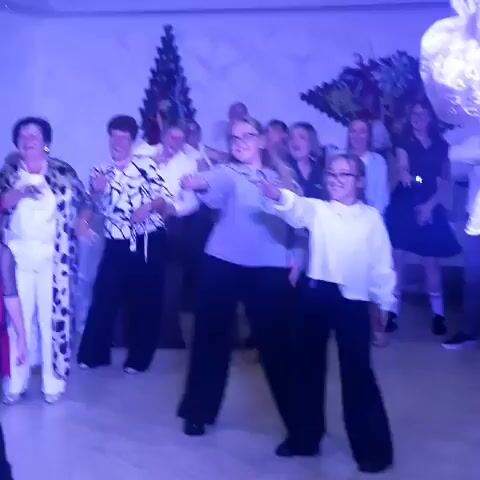 A group of people are dancing in a room with blue lighting. There are women of various ages, and a few men in the background. Two girls in the foreground are dancing enthusiastically, wearing glasses. One is in a white shirt and black pants, and the other is in a striped shirt and black pants. Other people in the background are also moving to the music. The room appears to be decorated with some sort of tree-like structures and foliage on the walls.