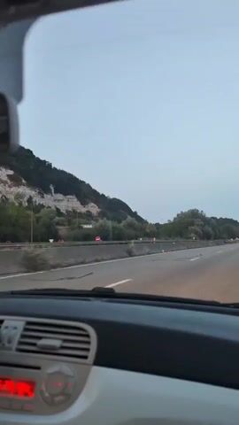 The video begins with a view from inside a car driving on a highway. The dashboard is visible in the foreground, with a portion of the air conditioning vents and a red light on the left. The road stretches ahead, bordered by a concrete barrier and then a hillside covered with trees and some rocky outcrops. As the car continues, a sign that says "SANER bienvenue" appears on the right side of the road. Further down the highway, a distinctive tall, spiral-shaped structure is visible in the distance, nestled amongst trees. The sky is a pale blue, indicating either early morning or late evening. The car is traveling at a steady pace on a multi-lane highway.