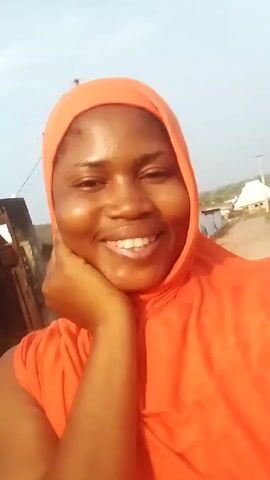 A person with dark skin and white teeth smiles at the camera. They are wearing an orange headscarf and shirt. They hold one hand up near their face. They are outside on a bright day. Behind them are buildings and power lines.