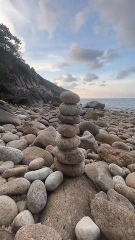 The video shows a cairn of eight flat, round rocks stacked on top of each other on a rocky beach. The beach is covered in similar rocks, and in the background is a hill with trees and the ocean. The sky is blue with some clouds.