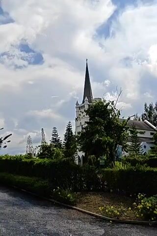 The video shows a panning shot of a church with a tall, pointed steeple. The church is white with dark trim and has large windows. In front of the church is a well-manicured garden with hedges and trees. A gravel driveway leads up to the church. The sky is blue with white clouds. There are two motorcycles parked near the church.