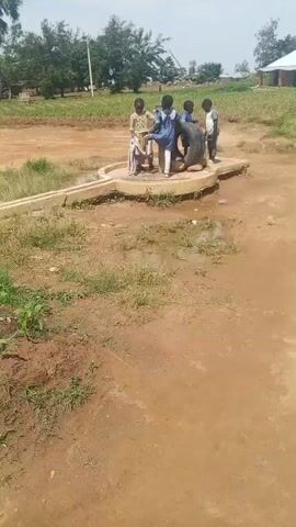 This media file shows four young children gathered around a large tire, which is placed on a concrete structure in an outdoor setting. The children appear to be playing with or interacting with the tire. They are all dressed in casual clothing.

The scene is set in a rural or semi-rural environment, with dirt ground, sparse grass, and some vegetation visible in the background, including trees. In the distance, there are some buildings, suggesting a small village or community. The sky is cloudy, indicating overcast weather. The time of day appears to be daytime, likely morning or afternoon.

The children's emotions are not clearly discernible due to the distance and image quality, but their posture suggests they are engaged in some form of play or activity. The concrete structure they are standing on seems to be part of a well or a water collection point, as there appears to be some water around it.

There is no visible text in the media file. The location context is Ohit, Nigeria.