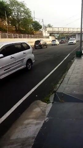 The video captures a street scene, likely during daylight, with a focus on a paved road and sidewalk. A silver minivan is prominently featured in the foreground, driving on the right side of the road. Several other cars, including a black SUV and white sedans, are visible further down the road, indicating moderate traffic. An overpass or bridge structure spans across the road in the background, with trees and greenery visible on the elevated embankment beside the road. The sidewalk appears to be paved with tiles, and there's a drainage grate or curb on the edge of the road. The overall environment suggests an urban or suburban setting. There are no people clearly visible or interacting in the scene. The camera appears to be stationary, looking towards the flow of traffic.