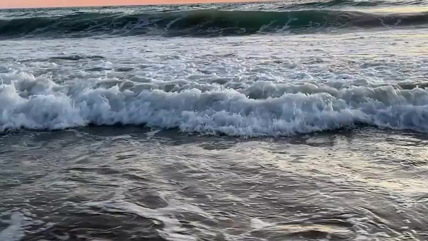 A serene ocean wave crashes onto a sandy shore during sunset. The sky displays a soft gradient of orange and pink, reflecting on the water's surface. The water is a mix of blues and grays, with white foam swirling as the wave recedes, leaving a trail of bubbles on the wet sand. The scene is tranquil and peaceful, with no people or objects visible.