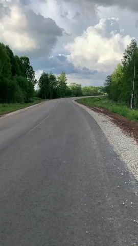 A paved road curves through a wooded area under a cloudy sky. The road surface appears to be asphalt, with a lighter colored gravel shoulder on the right side. Dense green trees line both sides of the road. The sky is filled with dramatic, white and gray clouds, suggesting the possibility of rain. The perspective is from a stationary point on the road, looking ahead as the road disappears over a gentle rise. There are no people or vehicles visible in the frame. The overall impression is one of a rural, somewhat overcast landscape.