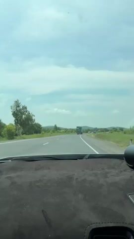 The video shows a view from inside a car looking out the windshield. The car is driving on a paved road with a white line marking the lanes. The road stretches into the distance, flanked by green grassy fields and trees. The sky is cloudy and appears to be daytime. In the distance, there are a few vehicles, including a truck. The dashboard of the car is visible at the bottom of the frame.