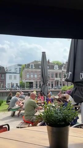 From under an awning, the scene shows a patio filled with people at picnic tables on a sunny day. Some tables are covered by large, gray umbrellas with the word “CORNET” printed on them. A small potted plant sits on the table from where the video is being shot.  In the background, across a canal, are buildings of various sizes and designs. People are sitting, standing, talking, eating, and drinking. A baby is crawling on the ground. Cars drive by on the street across the canal.
