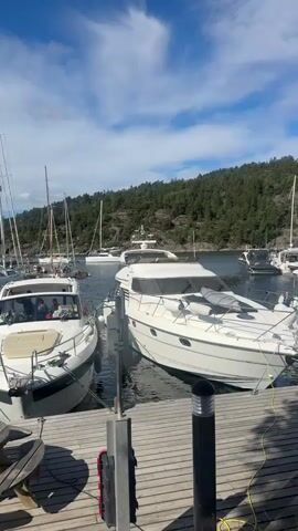 The video shows a series of boats docked in a marina on a sunny day. The boats are of various sizes and types, including yachts and sailboats. The marina is situated in a scenic location with a forested hillside in the background and a blue sky with scattered clouds overhead. The camera pans across the scene, offering views of the boats from different angles. There are no people visible in the video. The water appears calm and the overall atmosphere is peaceful.