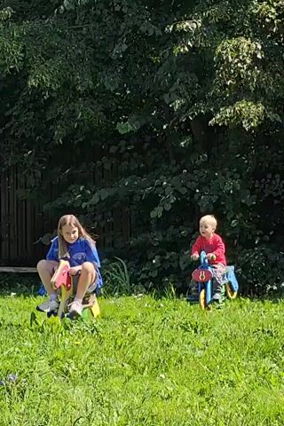 Two young children are playing on rocking toys in a grassy area with trees in the background. The child on the left, a girl with long blonde hair wearing a blue shirt and shorts, is actively rocking on a pink and yellow toy. The child on the right, a younger boy in a red shirt, is also on a blue and yellow toy, though his movements appear less vigorous. The sun is shining, creating bright light and shadows on the grass. A wooden fence is visible behind the girl.