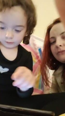 Sure, here is a concise description of the video:

The video captures a close-up shot of a young girl and a woman sitting side by side. The girl, who appears to be around three or four years old, has curly brown hair styled in pigtails. She is wearing a black long-sleeved shirt with a heart-shaped design. Her attention is focused on a smartphone in her hands, which she is tapping with her finger. Next to her sits a woman with long, red hair. She appears to be taking the video, holding the phone in front of them. The backdrop behind them includes a colorful bedspread or pillow with rainbow stripes. The lighting in the room is soft, and the overall focus is on the interaction between the child and the digital device, as well as the woman's presence.