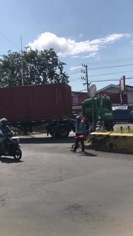 A large red shipping container truck is partially obstructing a road. A person wearing a high-visibility vest is directing traffic. Several motorcycles are present, with at least two riders visible. The environment appears to be a sunny outdoor location with trees and utility poles in the background. The camera is positioned at ground level, looking towards the truck and the traffic director.