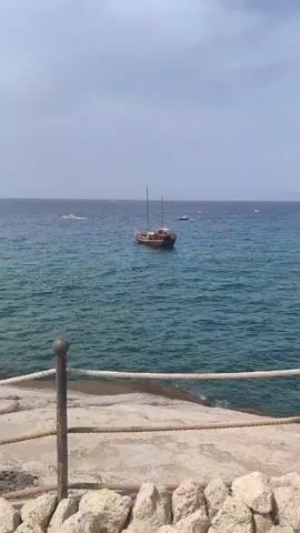 The video shows a view of the sea from a rocky shore. In the foreground, there is a stone pathway with a rope railing. The sea is a vibrant turquoise color, and there are several boats visible in the distance. The main focus is a wooden boat with two masts anchored in the water. It appears to be carrying passengers, as several people are visible on its deck. The sky is overcast, and the overall atmosphere is calm and serene.