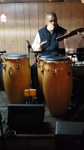 A man is playing two conga drums while standing in front of a wood-paneled wall. He is wearing a light-colored long-sleeved shirt and a dark vest. He is holding a striker in his right hand and using it to play a chimes instrument. The drums are a light tan color. There is a speaker on either side of the drums. The lighting is dim.