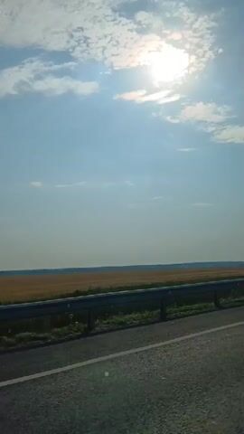 The video shows a view from a vehicle, looking out at a vast, golden field under a bright, sunny sky with scattered clouds. A highway guardrail runs horizontally across the frame, separating the road from the field. A tall, dark lamppost is visible on the right side of the frame. The perspective suggests the vehicle is moving, as the landscape appears to be shifting slightly. The sun is shining brightly, casting a glare in the upper portion of the sky. The overall scene depicts a rural landscape during daytime.