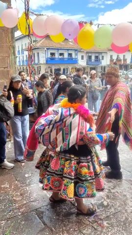 The video shows a group of people in a crowded outdoor space, likely a town square or plaza, under an archway decorated with colorful balloons. The people in the foreground are wearing traditional Andean clothing, including brightly colored embroidered skirts and shawls. They appear to be dancing or participating in a celebration. Others in the crowd, including some holding cell phones, watch the spectacle. The ground is wet and covered with confetti. Buildings surround the square, indicating an urban setting.