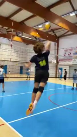 The video depicts a volleyball practice session in a gymnasium. Several young male players are on a blue court with white lines, separated by a net. One player, wearing a dark blue jersey with the number 14, is highlighted as he jumps and executes a volleyball hit. Other players can be seen around the court, some wearing light blue jerseys with the number 7. The gym has a wooden beam ceiling and white walls. A scoreboard is visible in the background, along with various sports-related posters and equipment. The camera follows the player with the number 14 as he moves on the court after his hit.