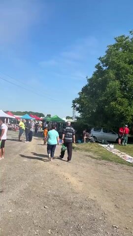 The video shows an outdoor market on a sunny day with a clear blue sky. People are browsing at stalls set up with colorful tents. Some individuals are walking and interacting with each other. Objects such as cars, bags, and bicycles are visible at some of the stalls, suggesting a flea market or car boot sale. The ground is dirt and gravel, and a large tree is present on the right side of the frame. There are power lines overhead. The atmosphere appears casual and active.